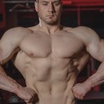 Intense shot of a muscular man posing topless in a gym setting showcasing physique.
